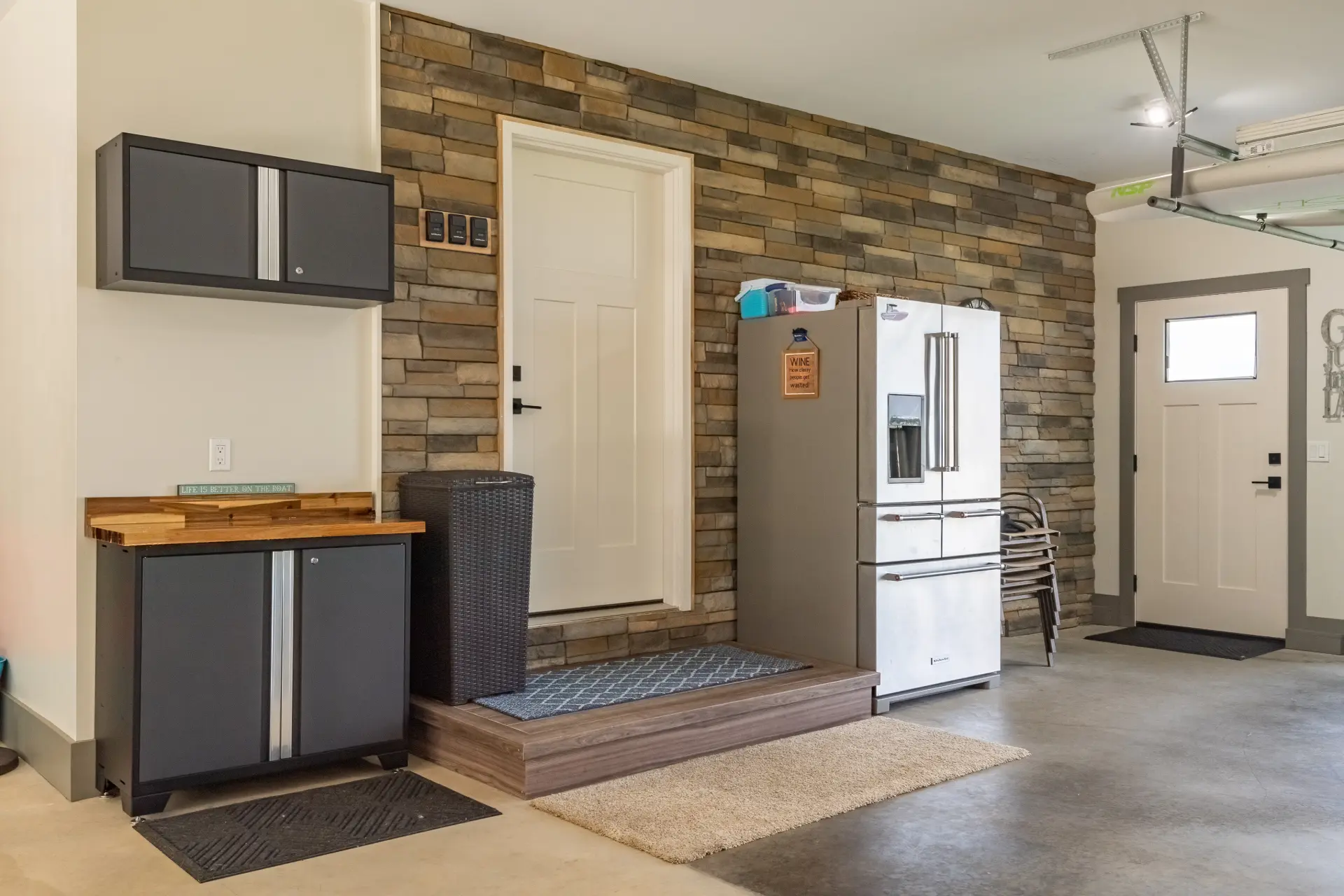 Finished garage entry area with stone accent wall, raised platform, exterior door, storage cabinets, refrigerator, and concrete floor