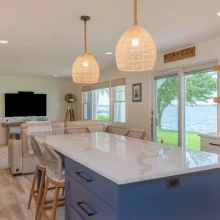 Open kitchen and living room with blue island, white countertops, woven pendant lighting, lake views, and bright waterfront windows