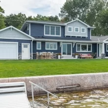Lakeside exterior of a Portage Austin Lake home with blue siding, covered porch, patio seating, green lawn, seawall, and dock access
