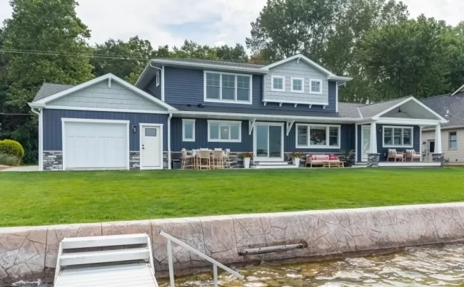 Portage Austin Lake Lakeside Exterior with Dock | Pennings & Sons Lakeside exterior of a Portage Austin Lake home with blue siding, covered porch, patio seating, green lawn, seawall, and dock access