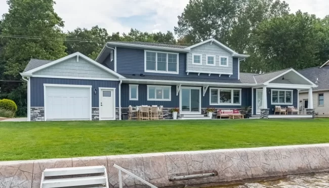 Lakeside exterior of a Portage Austin Lake home with blue siding, covered porch, patio seating, green lawn, seawall, and dock access