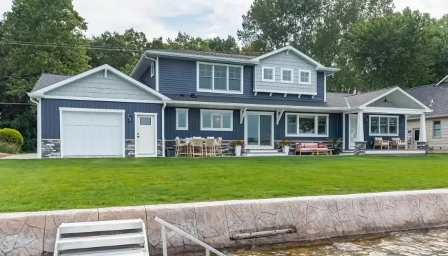 Portage Austin Lake Lakeside Exterior with Dock | Pennings & Sons Lakeside exterior of a Portage Austin Lake home with blue siding, covered porch, patio seating, green lawn, seawall, and dock access