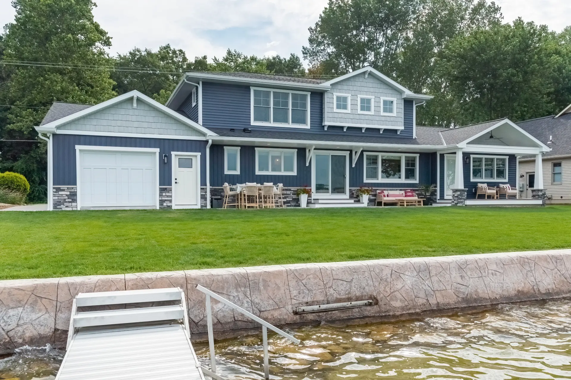 Lakeside exterior of a Portage Austin Lake home with blue siding, covered porch, patio seating, green lawn, seawall, and dock access