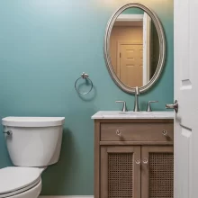 New light flooring that matches Kitchen in a light colored brick design, with small single sink vanity with oval mirror on wall above sink with white toilet to the left of the sink
