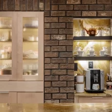 Built-in coffee bar set into a brick wall with lighted shelves, textured glass cabinet doors, coffee maker, cups, and stone countertop