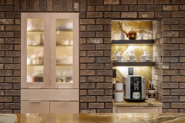 Built-in coffee bar set into a brick wall with lighted shelves, textured glass cabinet doors, coffee maker, cups, and stone countertop