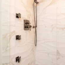 Custom shower with marble-look wall tile, handheld shower, rain showerhead, body sprays, black fixtures, and gray tile floor