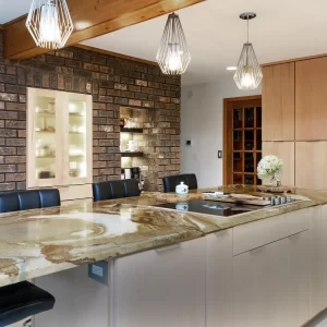 Kitchen island with natural stone countertop, brick accent wall, pendant lights, light wood cabinetry, and stainless steel double ovens