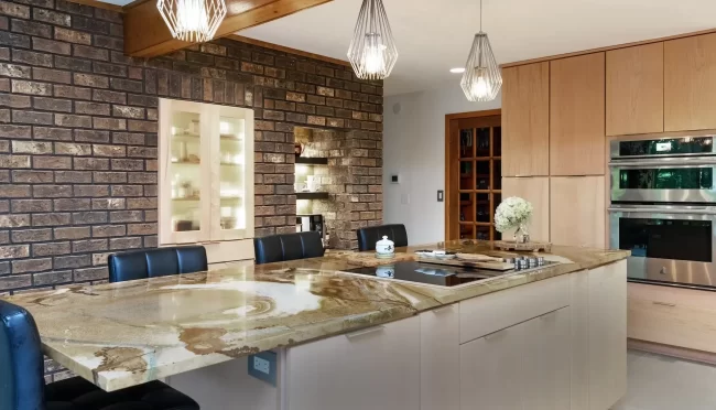 Kitchen island with natural stone countertop, brick accent wall, pendant lights, light wood cabinetry, and stainless steel double ovens
