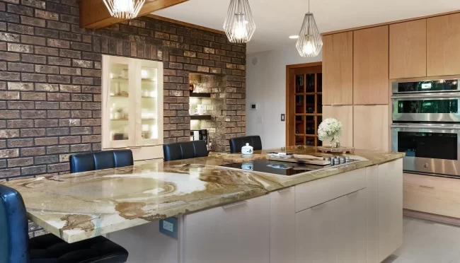 Kitchen island with natural stone countertop, brick accent wall, pendant lights, light wood cabinetry, and stainless steel double ovens