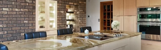 Kitchen Island with Brick Accent Wall | Pennings & Sons Kitchen island with natural stone countertop, brick accent wall, pendant lights, light wood cabinetry, and stainless steel double ovens