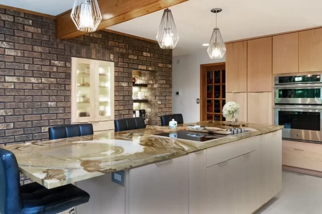 Kitchen Island with Brick Accent Wall | Pennings & Sons Kitchen island with natural stone countertop, brick accent wall, pendant lights, light wood cabinetry, and stainless steel double ovens
