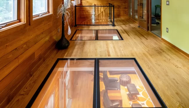 Loft Hallway with Glass Panels in Floor | Pennings & Sons Long wood-paneled hallway with glass floor panels, windows, decorative floor vase, and railing around the stair opening