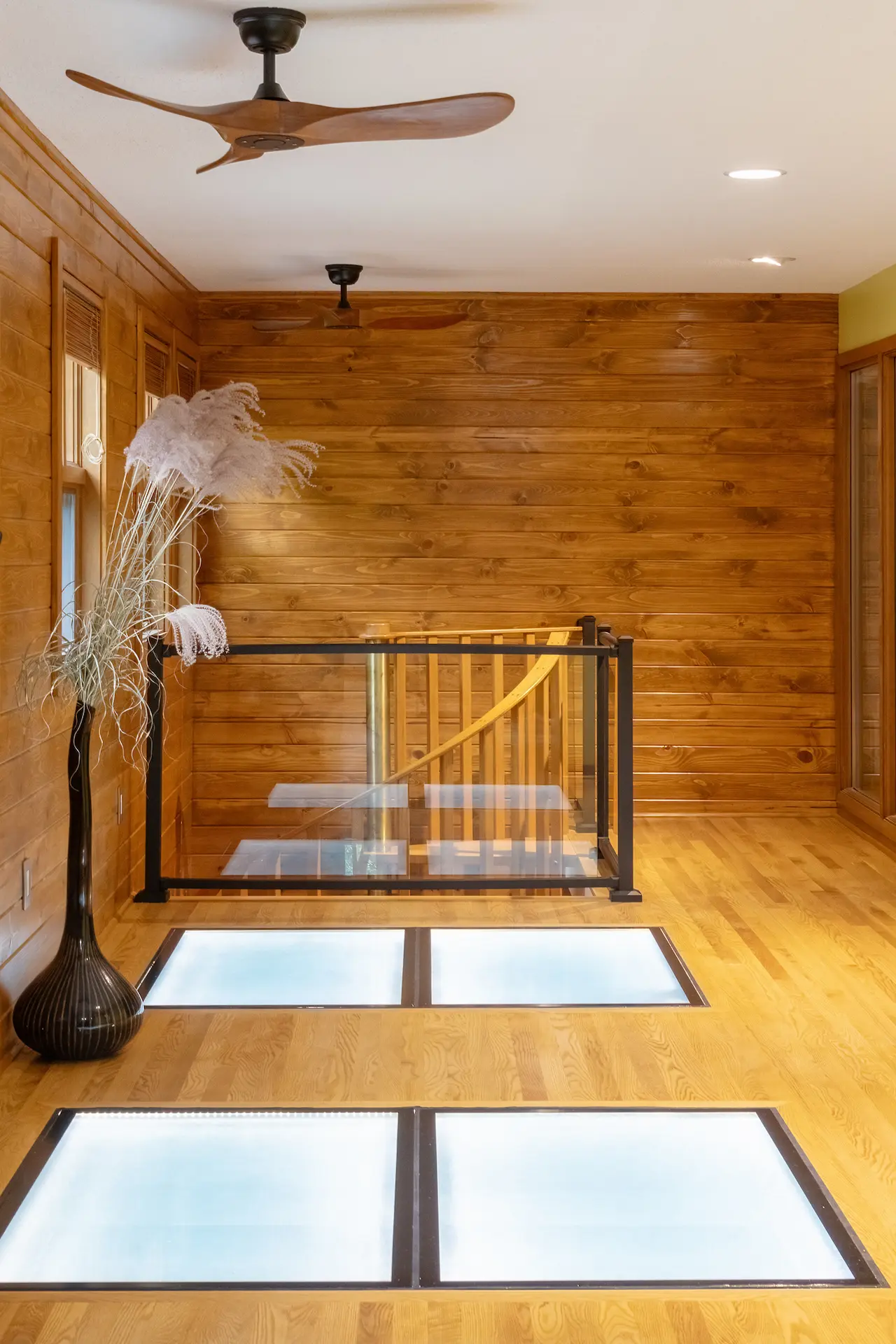 Loft landing with warm wood walls, wood flooring, glass floor panels, black railing, and spiral staircase below