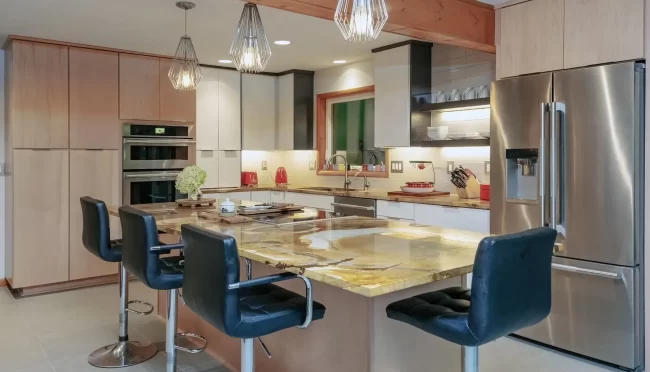 Modern kitchen with light wood cabinetry, large stone island, pendant lights, stainless steel refrigerator, and built-in appliances