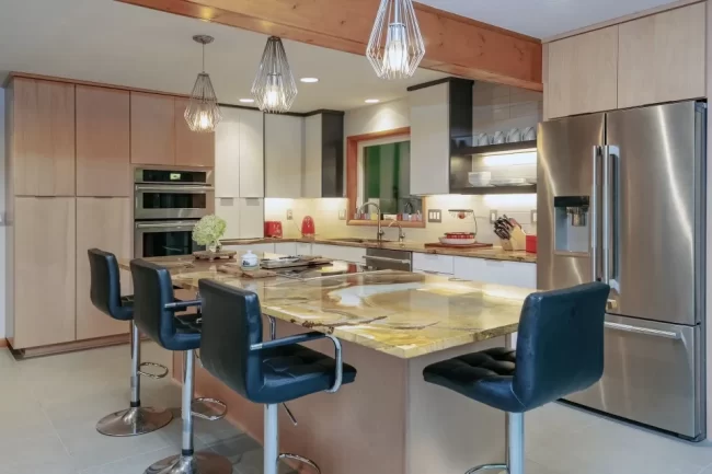 Modern kitchen with light wood cabinetry, large stone island, pendant lights, stainless steel refrigerator, and built-in appliances