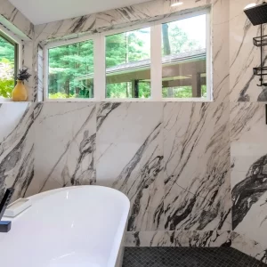 Soaking tub set against marble-look tile walls with black tub filler, tall windows, shower fixtures, and gray mosaic floor