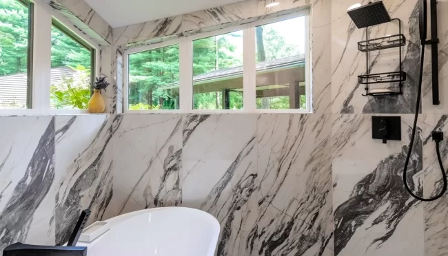 Southwest Michigan Soaking Tub and Marble-Look Tile Wall | Pennings & Sons Soaking tub set against marble-look tile walls with black tub filler, tall windows, shower fixtures, and gray mosaic floor