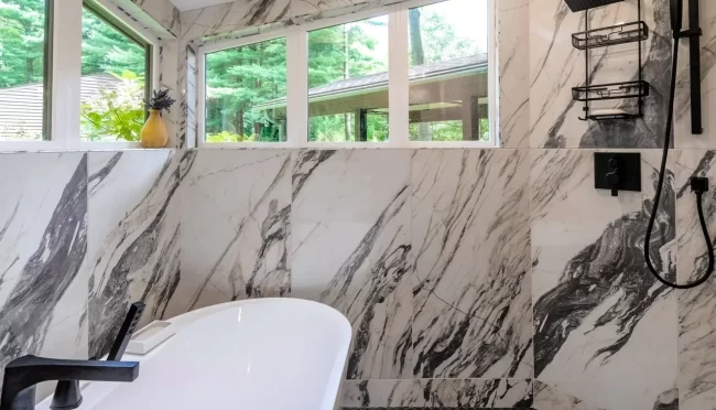 Soaking tub set against marble-look tile walls with black tub filler, tall windows, shower fixtures, and gray mosaic floor