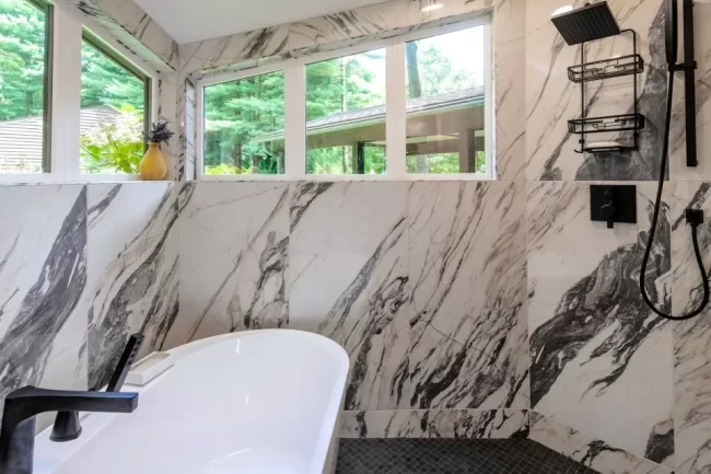 Soaking tub set against marble-look tile walls with black tub filler, tall windows, shower fixtures, and gray mosaic floor