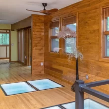 Wood-paneled hallway with illuminated glass floor panels, wood flooring, large windows, and black railing near the stair opening