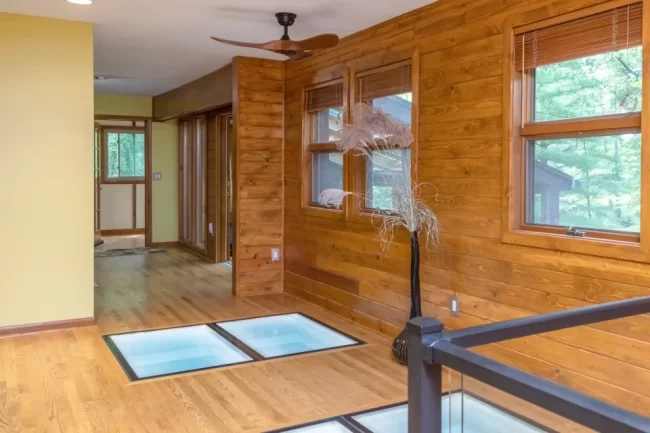Wood-paneled hallway with illuminated glass floor panels, wood flooring, large windows, and black railing near the stair opening
