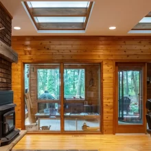 Wood-paneled living room with brick fireplace, wood stove, interior skylights, glass doors, and views to the screened porch