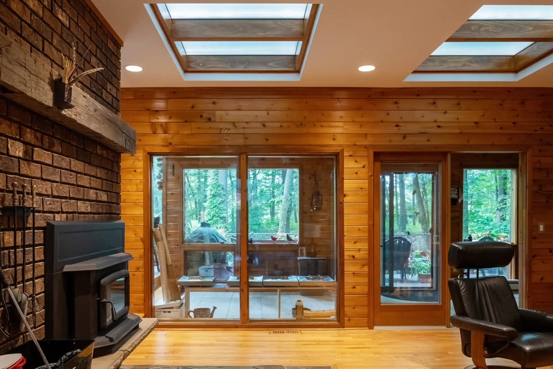 Wood-paneled living room with brick fireplace, wood stove, interior skylights, glass doors, and views to the screened porch
