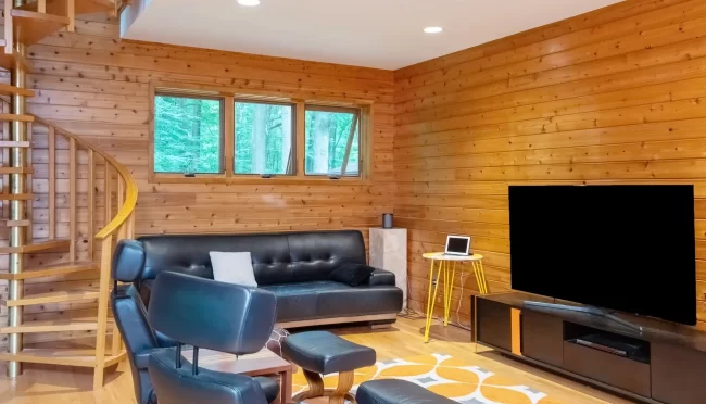 Wood Living Room with Spiral Staircase | Pennings & Sons Wood-paneled living room with spiral staircase, leather seating, wall-mounted TV, warm wood flooring, and large window