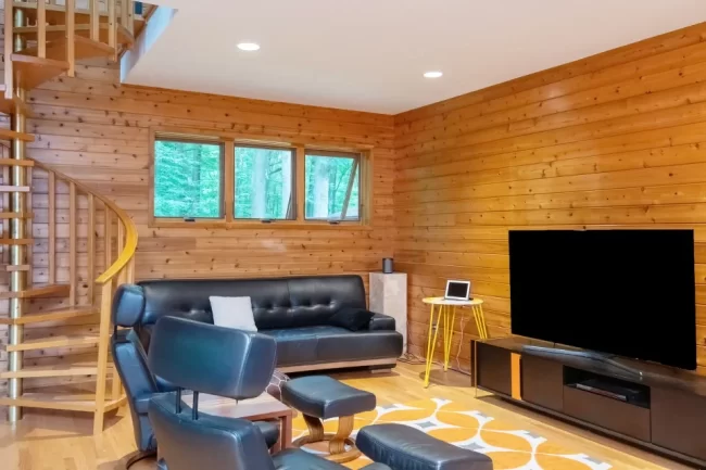Wood Living Room with Spiral Staircase | Pennings & Sons Wood-paneled living room with spiral staircase, leather seating, wall-mounted TV, warm wood flooring, and large window