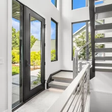 Modern entryway with black-framed glass door, floating stairs, cable railing, tall windows, and bright natural light