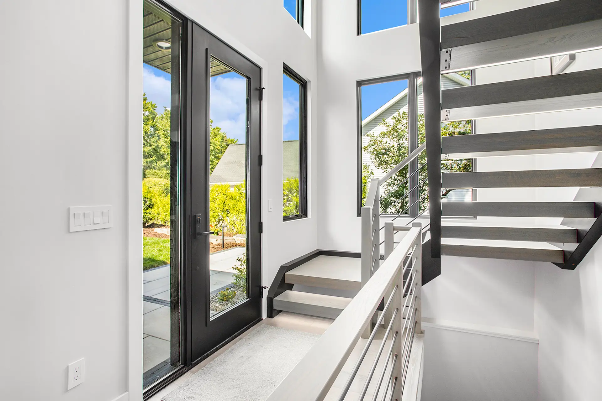 Modern entryway with black-framed glass door, floating stairs, cable railing, tall windows, and bright natural light