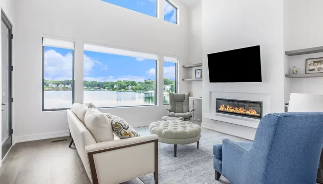 Crooked Lake Family Room Fireplace | Pennings & Sons Family room with fireplace, wall-mounted TV, built-in shelves, lake-facing windows, and comfortable seating.