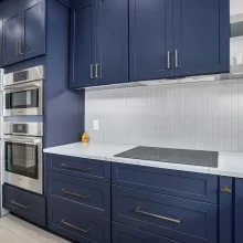 Custom kitchen with navy cabinets, stainless steel appliances, built-in ovens, white countertop, and tile backsplash.