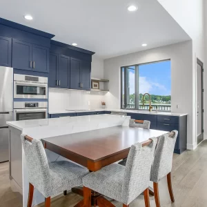 Custom kitchen with navy cabinetry, large white island, dining table extension, stainless appliances, and lake-facing windows.