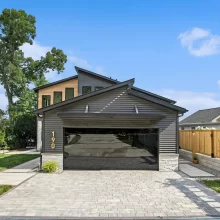 Modern lake home exterior with mirrored garage door, gray siding, stone accents, paver driveway, and landscaped front entry