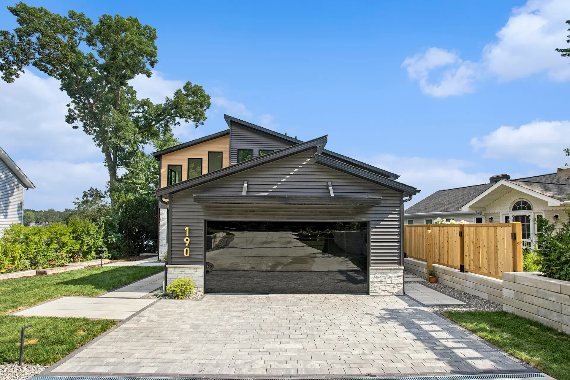 Modern lake home exterior with mirrored garage door, gray siding, stone accents, paver driveway, and landscaped front entry