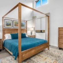 Primary bedroom with wood canopy bed, teal bedding, matching wood furniture, tall ceiling, large windows, and natural light