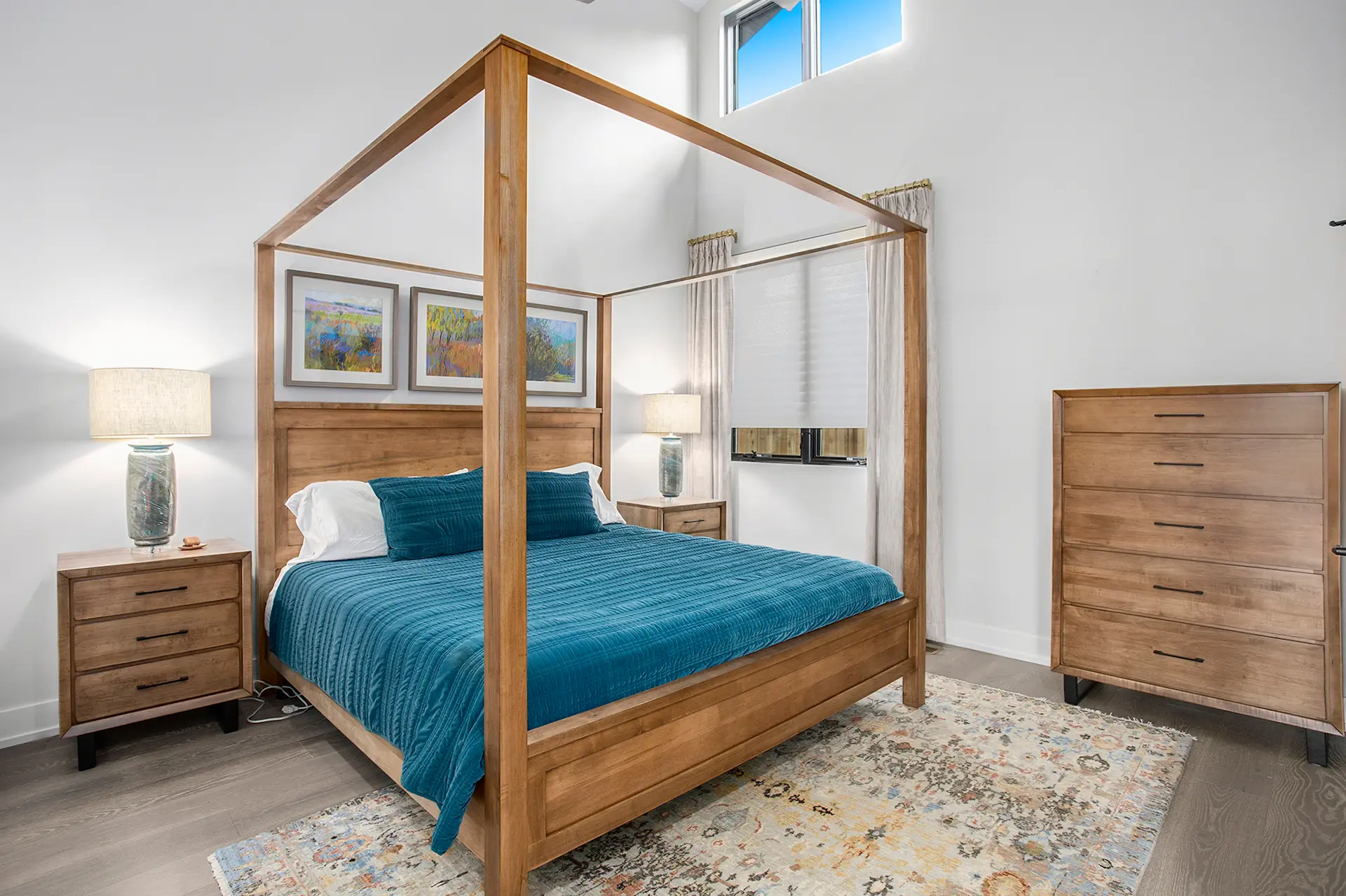 Primary bedroom with wood canopy bed, teal bedding, matching wood furniture, tall ceiling, large windows, and natural light