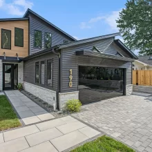 Street view of modern Crooked Lake home with mirrored garage door, gray siding, stone accents, paver driveway, and front walkway