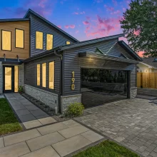 Twilight exterior view of modern Crooked Lake home with illuminated windows, mirrored garage door, paver driveway, and evening sky