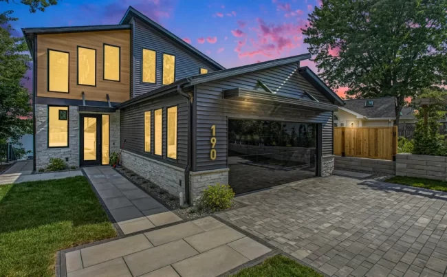 Crooked Lake Twilight Exterior | Pennings & Sons Twilight exterior view of modern Crooked Lake home with illuminated windows, mirrored garage door, paver driveway, and evening sky