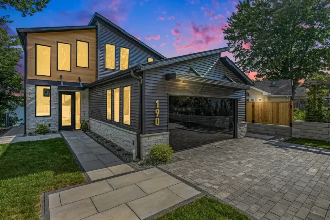 Crooked Lake Twilight Exterior | Pennings & Sons Twilight exterior view of modern Crooked Lake home with illuminated windows, mirrored garage door, paver driveway, and evening sky