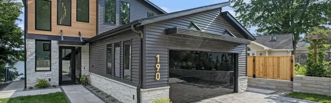 Crooked Lake Modern Home Street View | Pennings & Sons Street view of modern Crooked Lake home with mirrored garage door, gray siding, stone accents, paver driveway, and front walkway