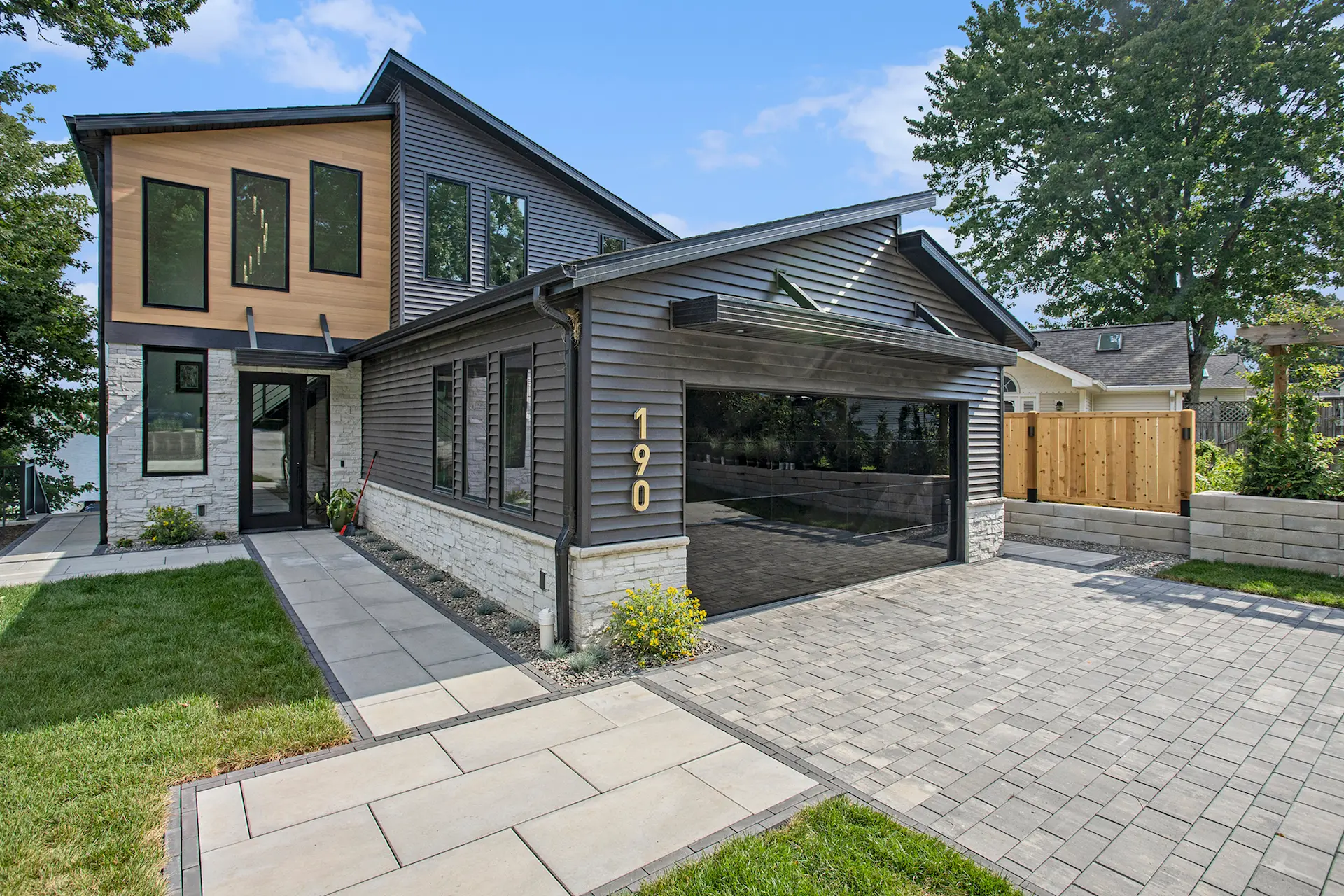 Street view of modern Crooked Lake home with mirrored garage door, gray siding, stone accents, paver driveway, and front walkway