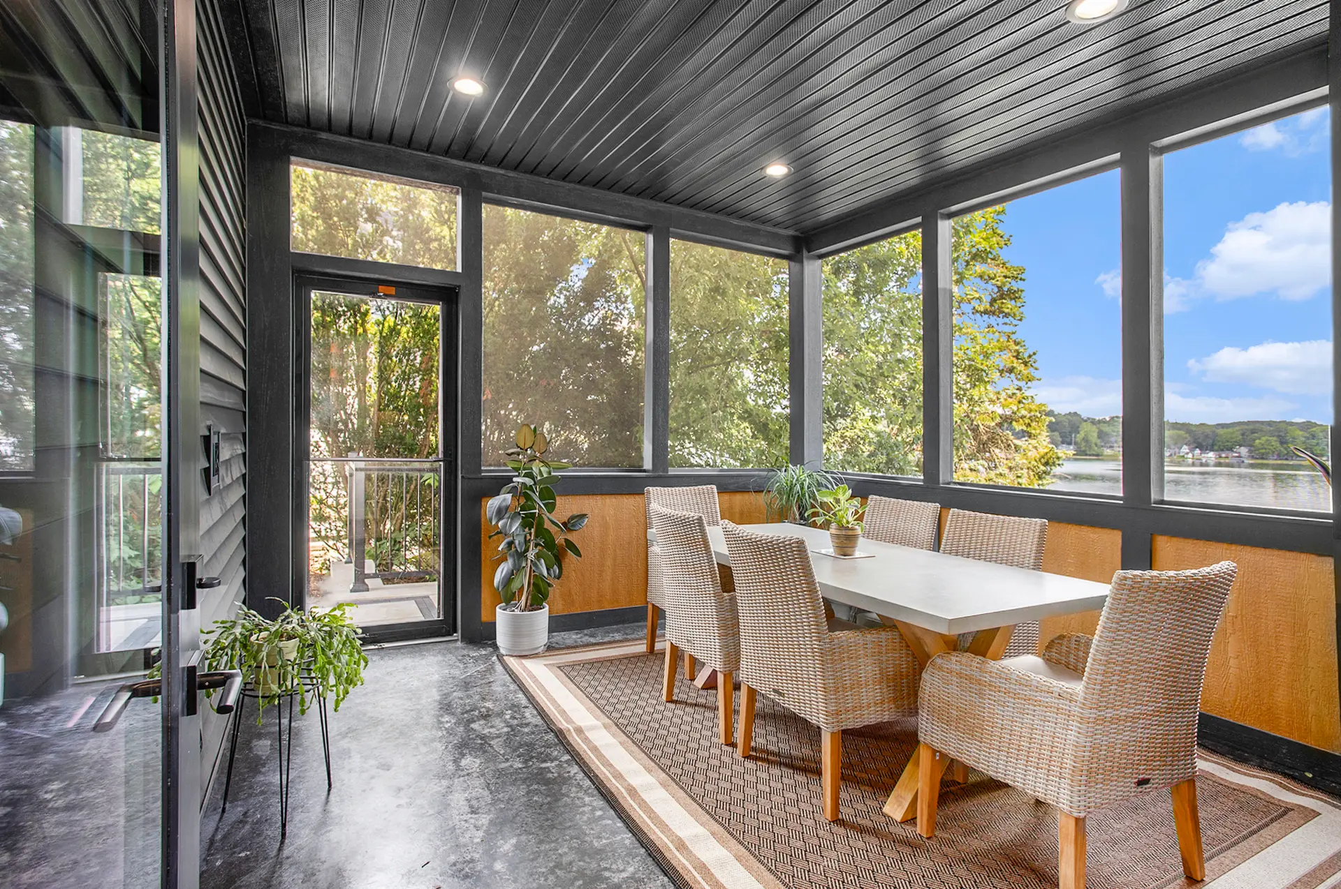 Three season porch with wicker dining chairs, black screened windows, wood accent walls, potted plants, and views of Crooked Lake