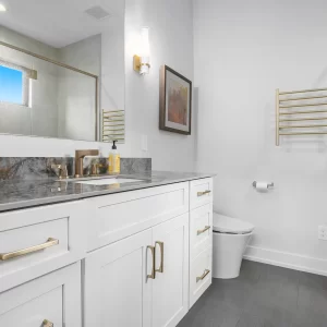 Primary bathroom with white vanity, gold hardware, stone countertop, wall mirror, gray tile floor, toilet, and glass shower