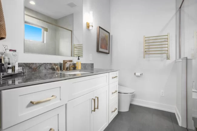 Crooked Lake Primary Bathroom | Pennings & Sons Primary bathroom with white vanity, gold hardware, stone countertop, wall mirror, gray tile floor, toilet, and glass shower