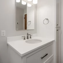 White bathroom vanity with white countertop, framed mirror, polished nickel faucet, vanity light, and tall linen cabinet