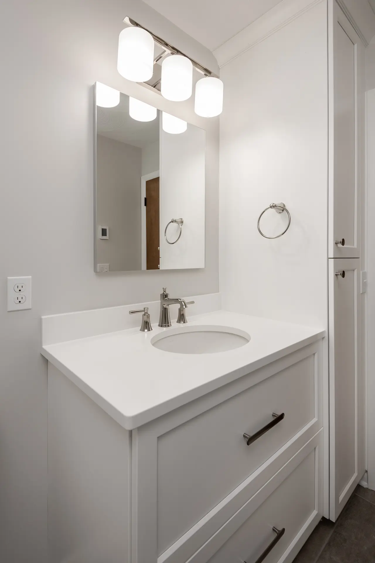 White bathroom vanity with white countertop, framed mirror, polished nickel faucet, vanity light, and tall linen cabinet